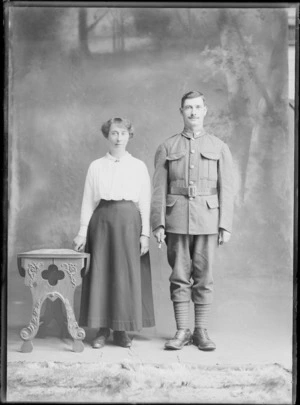 Studio unidentified family portrait of a young World War I soldier with moustache and fern collar badges, putties and hobnail boots standing holding a swagger stick next to his wife, Christchurch