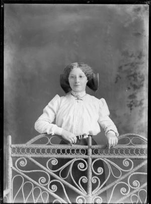 Studio unidentified portrait of a young woman with glasses and a large hair bow, in a high lace neck collar cotton shirt with greenstone bar and neck star jewelled brooches, chain and pearl pin, standing behind a couch, Christchurch