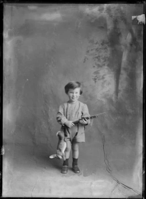 Studio unidentified family portrait of a young boy with a dragonfly embroidered jumpsuit holding a toy rifle with a rabbit hanging from his belt, Christchurch