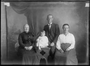 Studio unidentified family portrait, father with stiff wing shirt collar and striped tie, sitting behind toddler daughter in a lace dress on a highchair next to her mother and grandmother in a dark lace blouse and dress with greenstone bar neck brooch, Christchurch