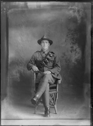 Studio portrait of an unidentified World War I soldier with bandolier ammunition belt over left shoulder, collar and hat badges and stirrups [Mounted Rifles?] sitting in a cane chair, Christchurch