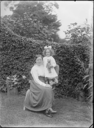 Outdoors on grass in front of a hedge, an unidentified family portrait of a mother with glasses sitting with her young daughters with hair bow standing holding a teddy bear, probably Christchurch region