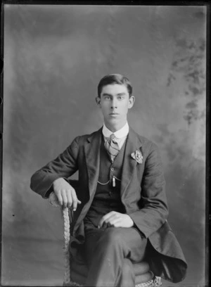 Studio unidentified portrait of a young man with double round shirt collar, striped tie, greenstone watch chain pendant and carnation, sitting in a cane chair, Christchurch