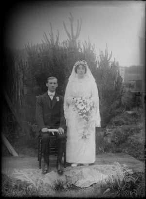Outdoors in backyard area in front of a pine tree, an unidentified wedding couple portrait, groom with white bow tie and lapel flowers sitting, bride with long veil holding flowers standing, probably Christchurch region