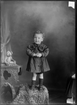Studio portrait of an unidentified small child, standing on a box with a teddy bear on table alongside, possibly Christchurch district