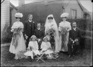 Wedding group, all members unidentified, including two small flowergirls wearing lace bonnets, sitting on an animal skin rug, holding staffs, in a garden, possibly Christchurch district