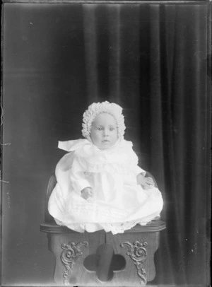 Studio portrait of an unidentified infant, wearing a gown and a ruched bonnet, sitting in a high wooden chair, possibly Christchurch district