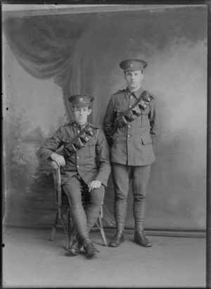 Studio portrait of two unidentified young men wearing military uniforms including gun cartridge holders worn across their chests, one man is sitting and the other standing, possibly Christchurch district