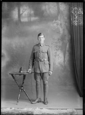 Studio portrait of unidentified World War I young soldier with collar and hat badges, standing holding a swagger stick with his hat on a table, Christchurch