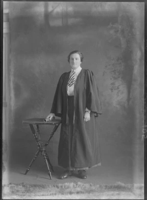 Studio portrait of an unidentified young woman in a graduation gown with a striped necktie standing holding a rolled up book next to a small table, Christchurch