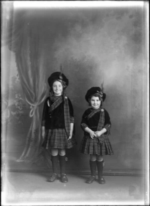Studio portrait of two unidentified girls in Scottish costume, wearing tartan kilts and sashes with brooches, large caps with clan crest pins and feathers, Christchurch