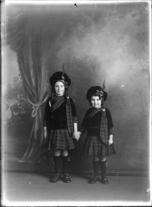 Studio portrait of two unidentified girls wearing tartan kilts and sashes with brooches, large caps with clan crest pins and feathers, Christchurch