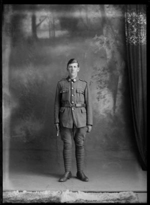 Studio portrait of unidentified World War I soldier with collar badges and black shoulder pocket cord, standing holding a swagger stick, Christchurch