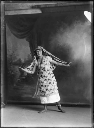 Studio portrait of unidentified young woman dressed as a silent movie actress, with bead necklaces and bracelets and matching patterned dress and headdress, Christchurch