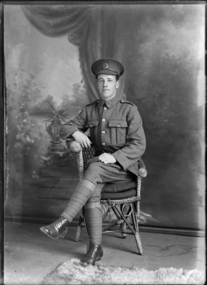 Studio portrait of unidentified World War I soldier sitting in a cane chair wearing his hat, with 'Liverpool' collar and hat badges [Rifle Brigade Reinforcements?], Christchurch