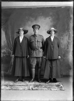 Studio portrait of unidentified World War I soldier with 'Liverpool' collar and hat badges [Rifle Brigade Reinforcements?], standing between two women in hats, Christchurch