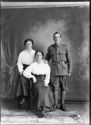 Studio portrait of unidentified World War I soldier with 'Liverpool' collar badges, standing beside two women with necklace and bar brooch, Christchurch