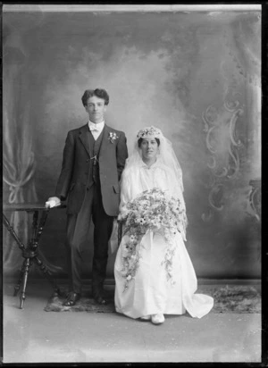 Studio unidentified wedding couple portrait, groom standing with white bow tie, buttonhole and pocket watch chain greenstone tiki, bride sitting with long veil holding flowers, Christchurch