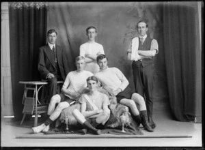 Studio portrait of unidentified bicycle road racing team, four young men in riding attire with two small cups, manager on left with polka dot tie and coach on right, Christchurch