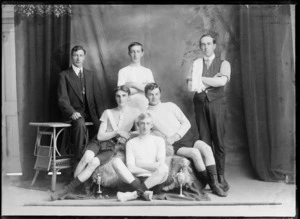 Studio portrait of unidentified bicycle road racing team, with four young men in riding attire in front with two small cups, manager with polka dot tie and coach alongside, Christchurch