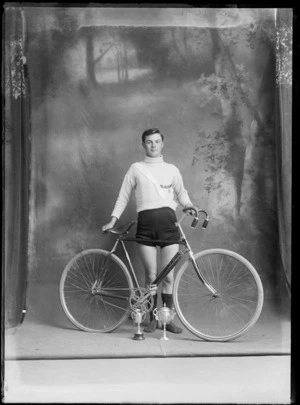 Studio portrait of unidentified bicycle road racing team member in riding attire with sash and two medals, holding his bike with two small cups in front, Christchurch