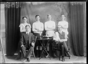 Studio portrait of unidentified bicycle road racing team, with four young men in riding attire standing, manager with polka dot tie and coach in front sitting beside table with small cups and medals, Christchurch