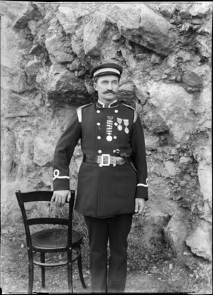 Outdoors portrait of unidentified man with a handlebar moustache in dress uniform with braided collar and hat, sleeve insignia and metal shoulder epaulettes, three lapel medals and a metal tool held in his belt, probably Christchurch region