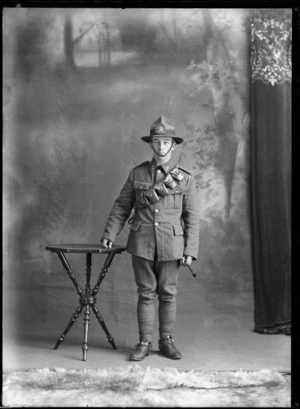 Studio portrait of unidentified World War I soldier with lemon squeezer hat, collar and hat badges, with bandolier ammunition belt over left shoulder, and stirrups and riding crop in hand [Mounted Rifles?], Christchurch