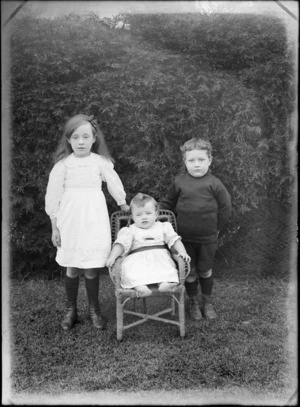 Portrait of three unidentified children taken outdoors, young girl in a lace dress and boy standing, baby with a lace bib and greenstone bar brooch in a cane chair, probably Christchurch region