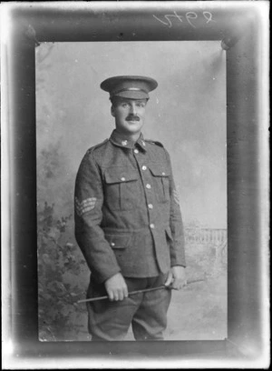 Studio portrait of unidentified World War I soldier with moustache, collar badges and hat, standing holding a swagger stick, probably Christchurch region