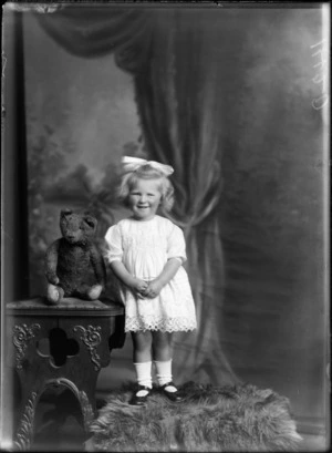 Studio portrait of unidentified young girl in lace dress with hair bow standing on a fur rug next to a large teddy bear, Christchurch
