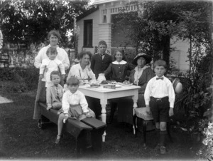 Group of women and children, in a garden, around a table laid out with afternoon tea items