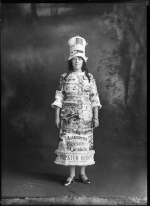 Studio portrait of unidentified young woman in a fancy dress and hat made from Aulsebrook's products advertising signs, Christchurch
