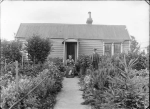 Unidentified elderly couple, with caged parrot and dog, in their front vegetable garden with single story wooden house behind, probably Christchurch region