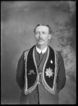 Studio portrait of unidentified older man with ornate frilly collar sash, with writing in Latin around border and 'PA' embroidered and star medallion and shield medal attached, [Hibernian Australasian Catholics Benefit Society members?], Christchurch