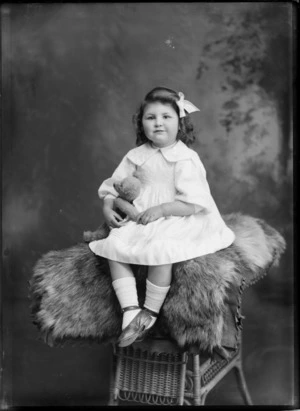 Studio portrait of an unidentified young girl with bow in hair and holding teddy bear, sitting on top of fur covered cane chair, Christchurch