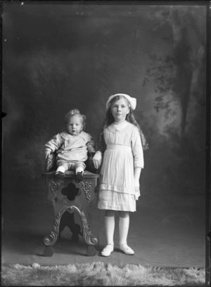 Studio portrait of unidentified young girl with long hair wearing a hat and gloves, standing next to a young baby on high chair, Christchurch