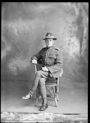 Studio portrait of unidentified young World War One soldier and hat with white band and badge, sitting in cane chair with riding crop in hand, wearing putties and stirrups [Mounted Rifles?], Christchurch