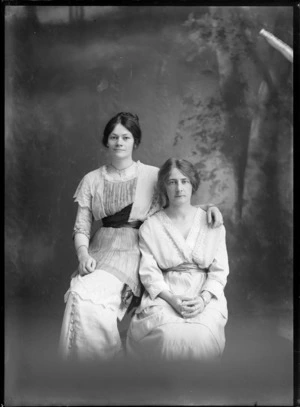 Studio portrait of two unidentified women in lace dresses with cummerbunds, bracelets and necklace, sharing a chair, Christchurch