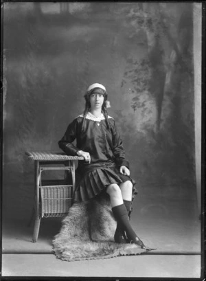Studio portrait of an unidentified young woman with long curly hair and ribbon, in a dark shirt and matching pleated skirt with large lace collar, heart shaped locket and bracelet, sitting with a cane table, Christchurch