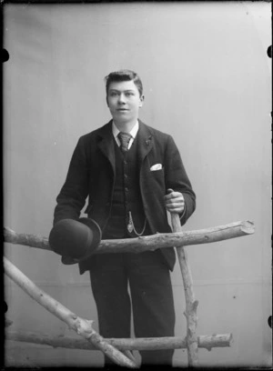 Outdoors portrait of unidentified young man in a three piece suit, bowler hat with a pocket watch chain pendant, standing with a prop wooden fence, probably Christchurch region