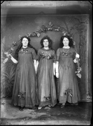 Outdoors portrait of three unidentified young women standing in costumes with berries and leaves, a star pendant and white elbow gloves, holding a leaf encrusted arched branch, probably Christchurch region