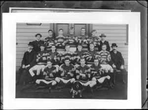Unidentified rugby football team portrait taken in front of wooden building, with fifteen players in uniform, two coaches with bowler hats and large moustaches, and a Cocker Spaniel dog, probably Christchurch region