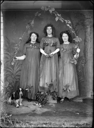 Outdoors portrait of three unidentified young women in costumes, with berries and leaves, a star pendant and white elbow gloves, holding a leaf encrusted arched branch, beside a Cocker Spaniel dog, probably Christchurch region
