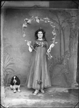 Outdoors portrait of unidentified young woman in a costume with berries and leaves, a star pendant and white elbow gloves, holding a leaf encrusted arched branch, beside a Cocker Spaniel dog, probably Christchurch region