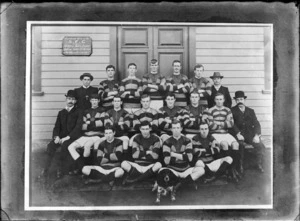 Unidentified rugby football team portrait in front of wooden building with 'SFC, H C Harrison' sign, includes fifteen players in uniform with four men in suits and a Cocker Spaniel dog, probably Christchurch region
