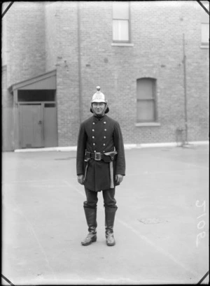 Outdoors portrait of unidentified young man in fire brigade uniform with a metal helmet, leather boots and belt equipment, [Christchurch Fire Brigade?], standing in front of brick building