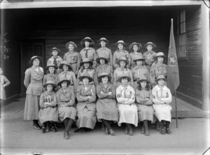 Outdoors portrait of the Sydenham Girl Scouts Troop with twenty one unidentified young women and one adult den mother troop leader with merit badges and company flag, Sydenham, Christchurch