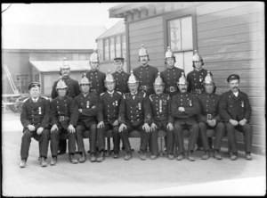 Outdoors in front of a large wooden building, a group portrait of fifteen unidentified Christchurch Regional Fire Brigade members in full dress uniforms with crested helmets and service medals, Christchurch region