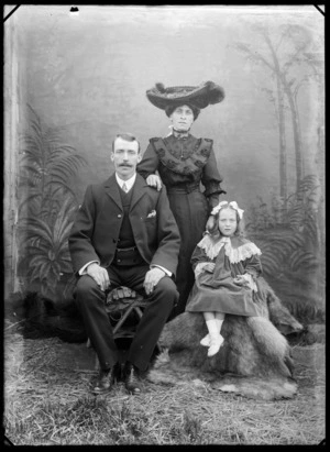 Outdoors portrait of unidentified family in front of a false backdrop, father with large moustache sitting next to young daughter in a lace fringed felt dress with hair bows, the mother in a dark flower embroidered patterned dress with greenstone bar brooch and large hat standing behind, probably Christchurch region